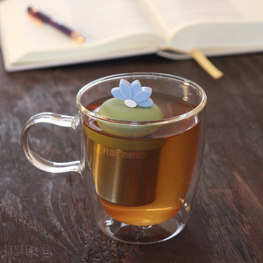 blue lotus flower tea infuser for loose leaf tea  with a fine mesh stainless steel tea infuser that reads FLOAT2INFUSE on a green base floating in a glass tea mug with notebook and pen in the background