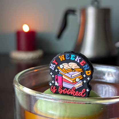 black round background with text "my weekend is booked" with a stack of books and a water bottle tea infuser  for loose leaf tea with fine mesh stainless steel tea strainer that reads FLOAT2INFUSE on a green base with a lit candle and a tea kettle in the background