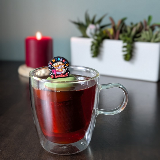 black round background with text "my weekend is booked" with a stack of books and a water bottle tea infuser  for loose leaf tea with fine mesh stainless steel tea strainer that reads FLOAT2INFUSE on a green base with a lit candle and plants in the background