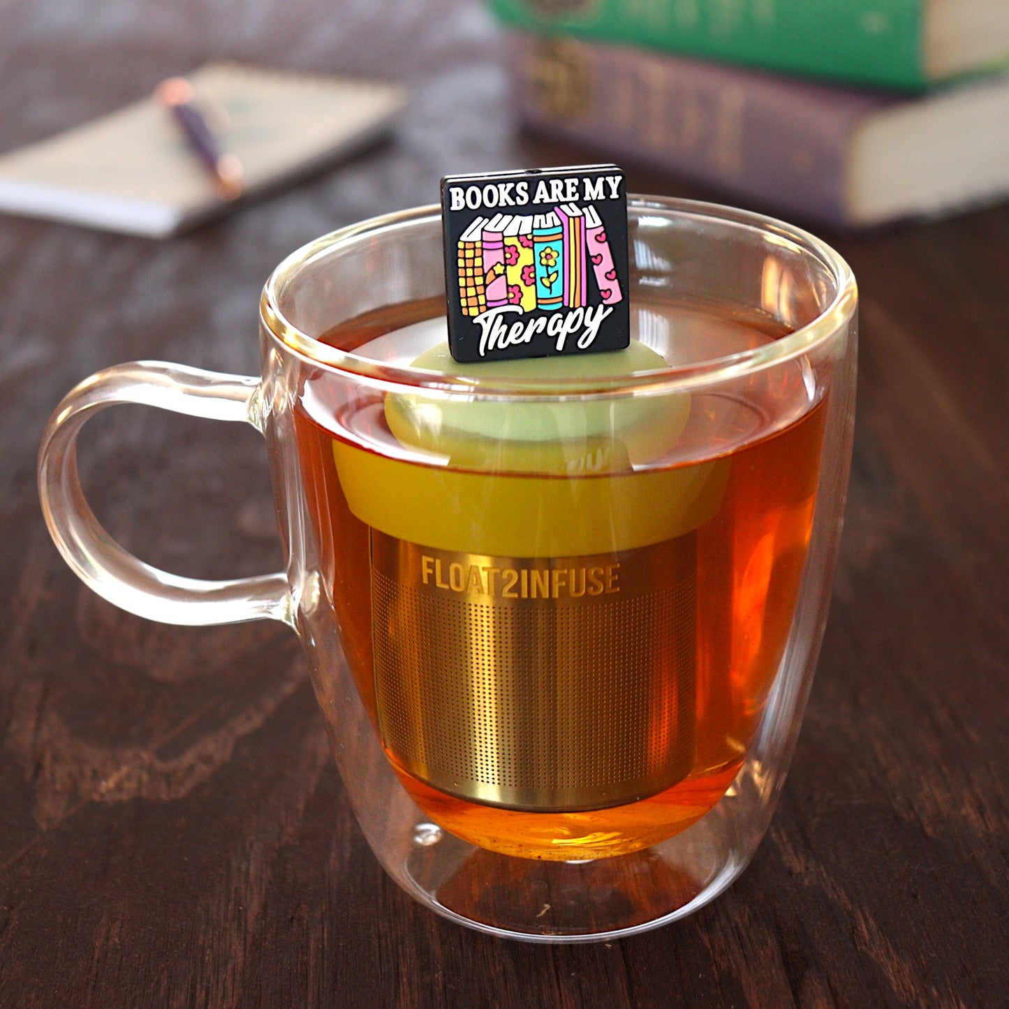 black square with text that reads "books are my therapy" on books tea infuser for loose leaf tea  with a fine mesh stainless steel tea infuser that reads FLOAT2INFUSE on a green base floating in a glass tea mug with pen on notebook and books in the background