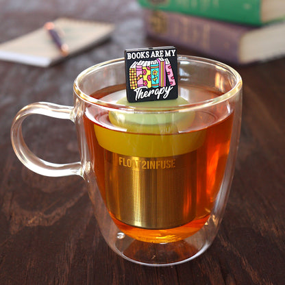 black square with text that reads "books are my therapy" on books tea infuser for loose leaf tea  with a fine mesh stainless steel tea infuser that reads FLOAT2INFUSE on a green base floating in a glass tea mug with pen on notebook and books in the background