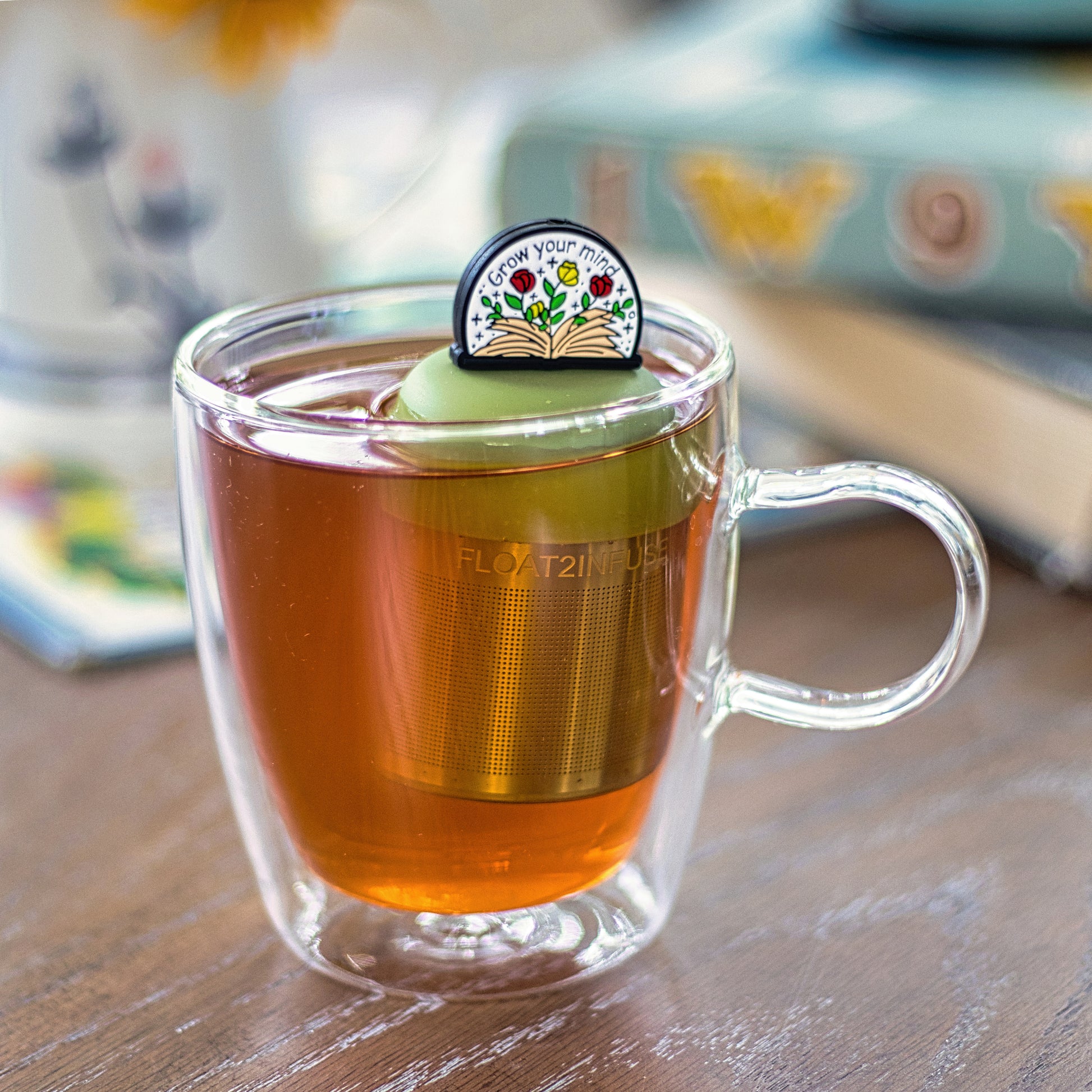 open book with flowers and text "grow your mind" tea infuser for loose leaf tea with a fine mesh stainless steel tea strainer that reads FLOAT2INFUSE on a green base  floating in a glass tea mug 
with books in the background