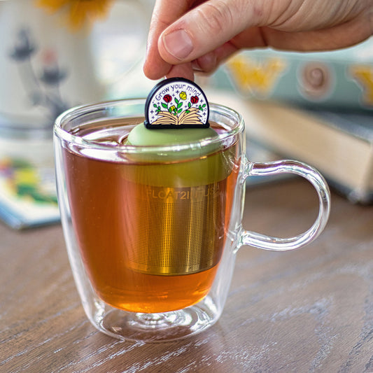 open book with flowers and text "grow your mind" tea infuser for loose leaf tea with a fine mesh stainless steel tea strainer that reads FLOAT2INFUSE on a green base  floating in a glass tea mug 
with books in the background