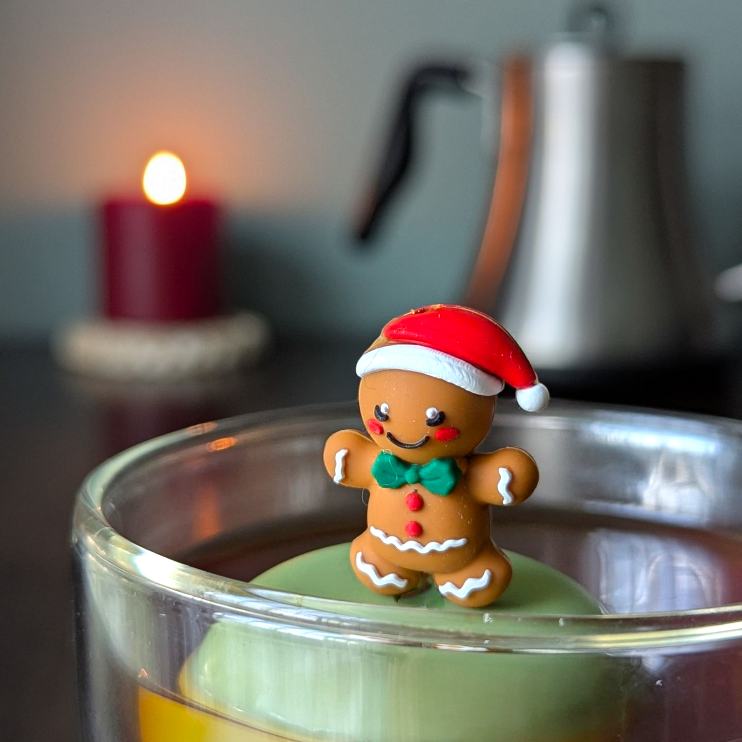 Smiling gingerbread man wearing a red santa hat and a green bowtie infuser for loose leaf tea with a fine mesh stainless steel tea strainer that reads FLOAT2INFUSE on a green base  with  a lit candle and a tea kettle in the background