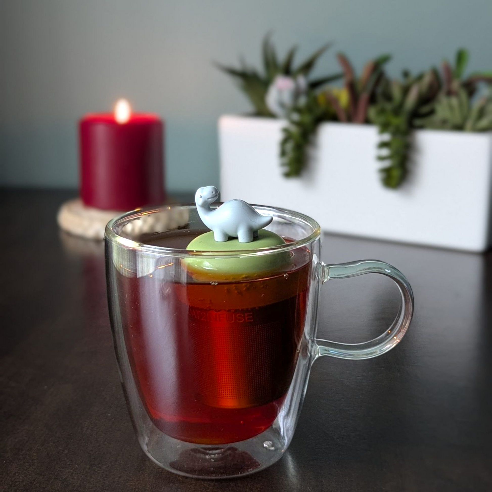 smiling grey brachiosaurus dinosaur tea infuser  for loose leaf tea with fine mesh stainless steel tea strainer that reads FLOAT2INFUSE on a green base with a lit candle and plants in the background