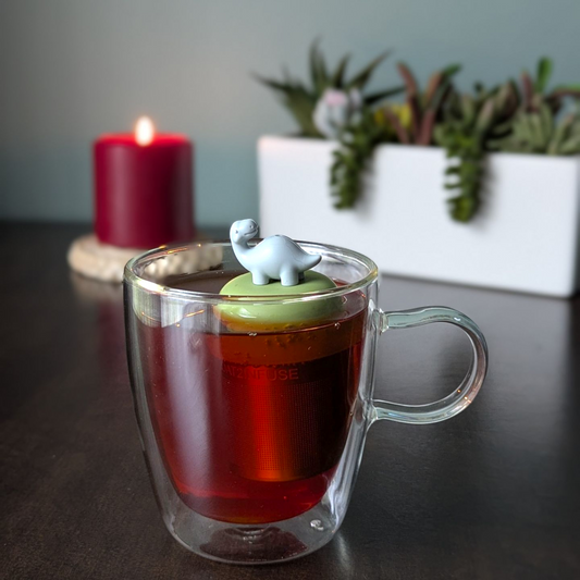 smiling grey brachiosaurus dinosaur tea infuser  for loose leaf tea with fine mesh stainless steel tea strainer that reads FLOAT2INFUSE on a green base with a lit candle and plants in the background