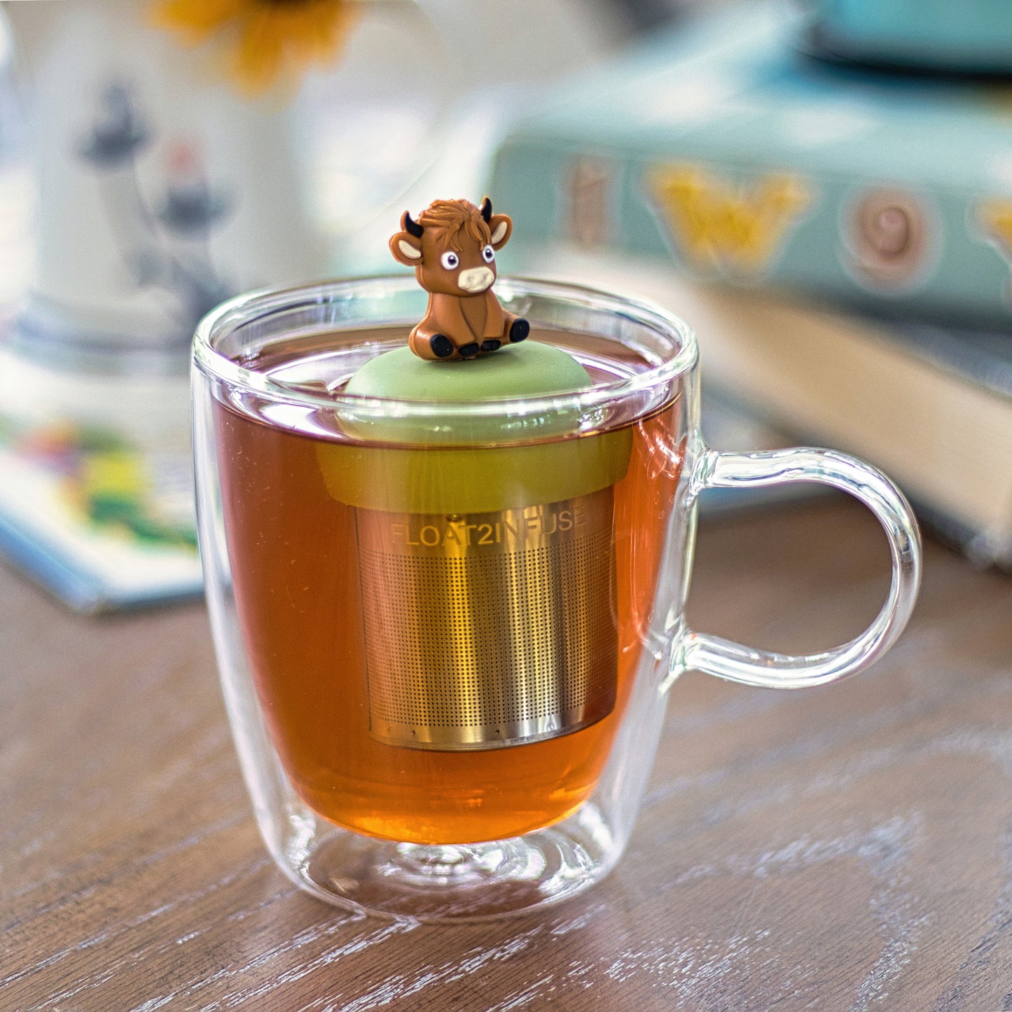 baby brown highland cow animal tea infuser for loose leaf tea with a fine mesh stainless steel tea strainer that reads FLOAT2INFUSE on a green base  floating in a glass tea mug with books in the background