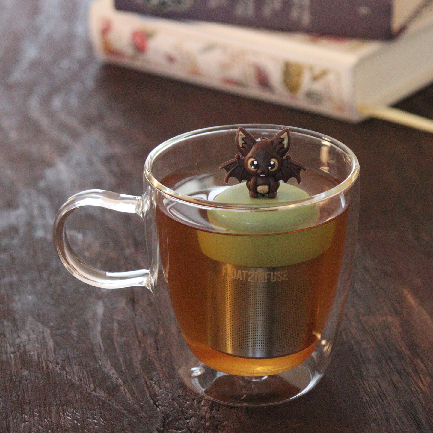 brown bat animal tea infuser for loose leaf tea  with a fine mesh stainless steel tea infuser that reads FLOAT2INFUSE on a green base floating in a glass tea mug with books in the background