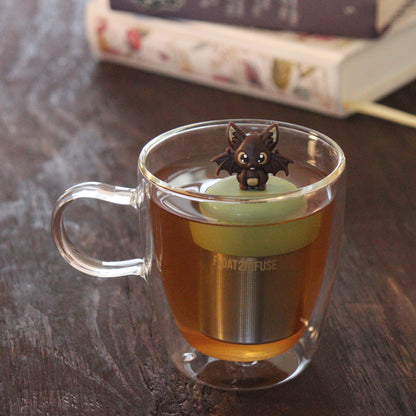 brown bat animal tea infuser for loose leaf tea  with a fine mesh stainless steel tea infuser that reads FLOAT2INFUSE on a green base floating in a glass tea mug with books in the background