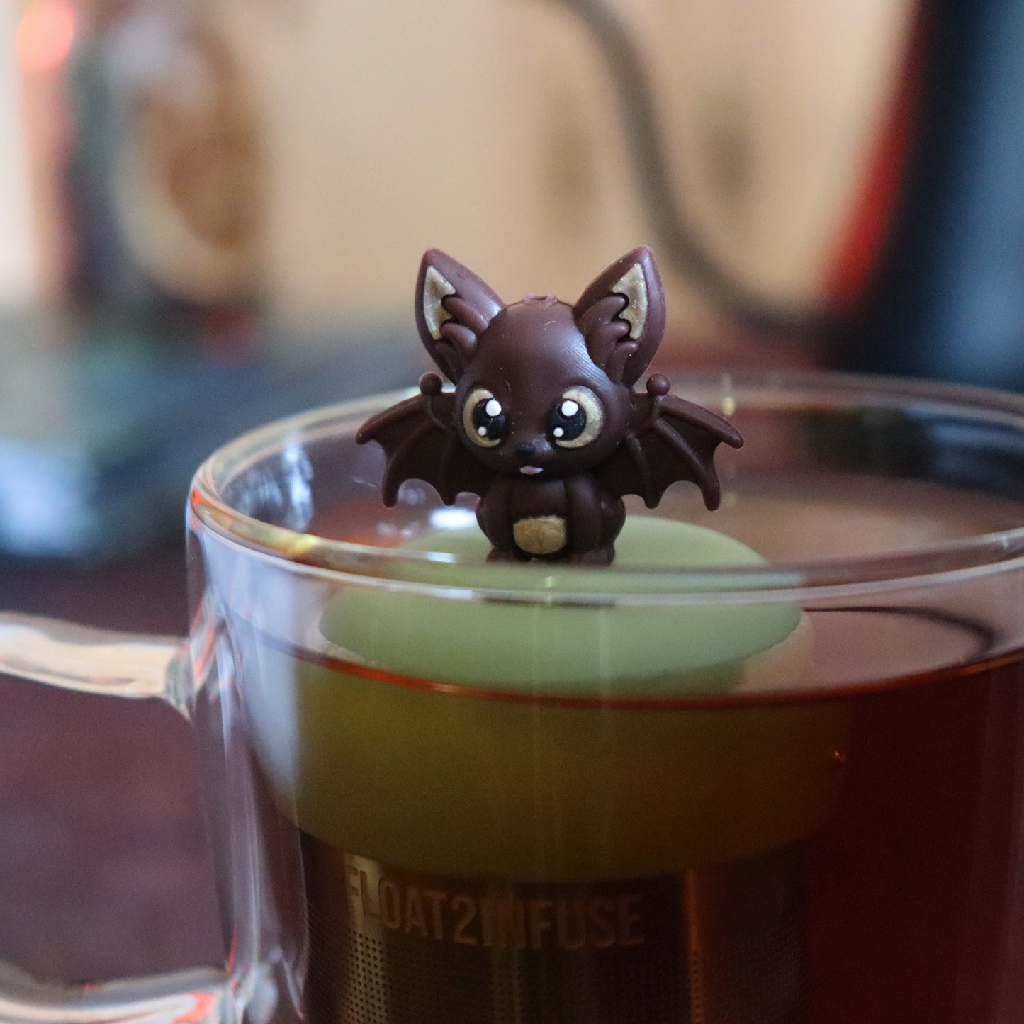 brown bat animal tea infuser for loose leaf tea with a fine mesh stainless steel tea strainer that reads FLOAT2INFUSE on a green base floating in a glass tea mug with tea kettle in the background