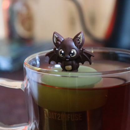 brown bat animal tea infuser for loose leaf tea with a fine mesh stainless steel tea strainer that reads FLOAT2INFUSE on a green base floating in a glass tea mug with tea kettle in the background