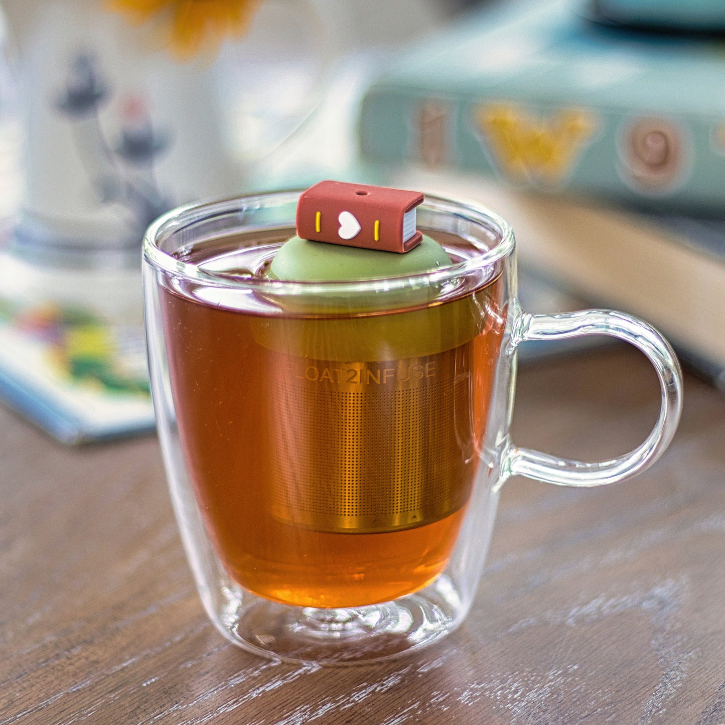 brown book with a white heart tea infuser for loose leaf tea with a fine mesh stainless steel tea strainer that reads FLOAT2INFUSE on a green base  floating in a glass tea mug with books in the background
