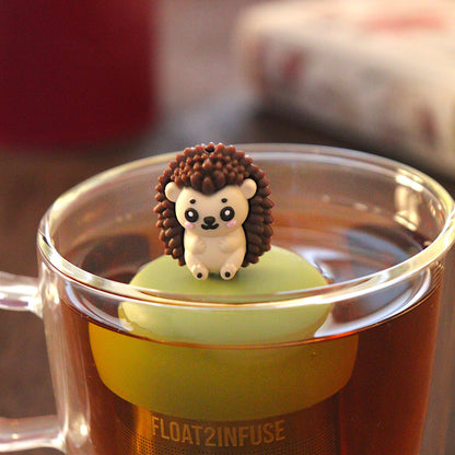 brown hedgehog tea infuser for loose leaf tea  with a fine mesh stainless steel tea infuser that reads FLOAT2INFUSE on a green base floating in a glass tea mug with loose leaf tea and books in the background