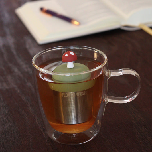 brown mushroom tea infuser for loose leaf tea  with a fine mesh stainless steel tea infuser that reads FLOAT2INFUSE on a green base floating in a glass tea mug with notebook and pen in the background