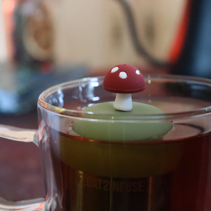 brown mushroom tea infuser for loose leaf tea with a fine mesh stainless steel tea strainer that reads FLOAT2INFUSE on a green base  floating in a glass tea mug with tea kettle in the background