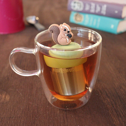 Brown squirrel holding an acorn tea infuser for loose leaf tea with a fine mesh stainless steel tea strainer 
that reads FLOAT2INFUSE on a green base with a tea canister and books in the background
