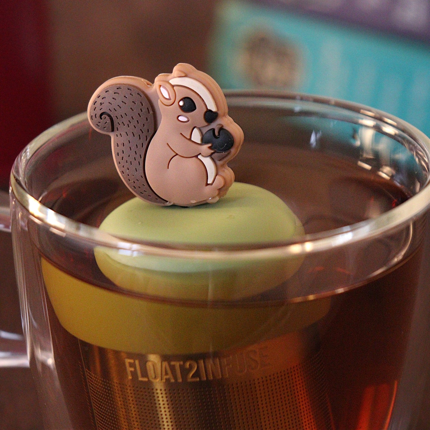 Brown squirrel holding an acorn tea infuser for loose leaf tea with a fine mesh stainless steel tea strainer 
that reads FLOAT2INFUSE on a green base with a tea canister and books in the background