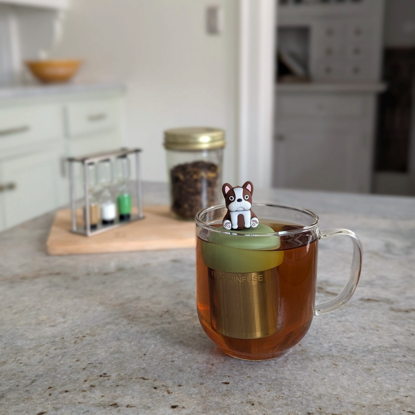 brown and white terrier dog tea infuser  for loose leaf tea with fine mesh stainless steel tea strainer that reads FLOAT2INFUSE on a green base floating in a glass tea mug with loose leaf tea in the background
