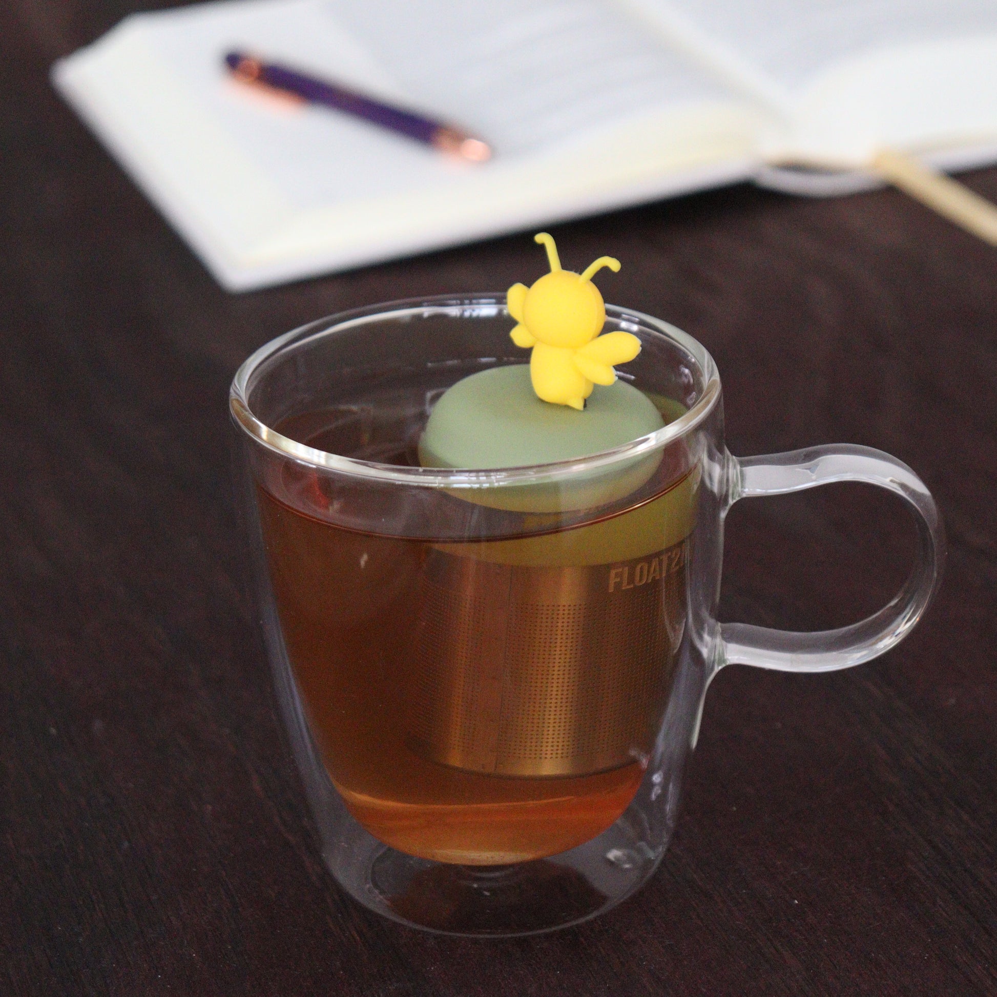 backside of yellow bee animal tea infuser for loose leaf tea  with a fine mesh stainless steel tea infuser that reads FLOAT2INFUSE on a green base floating in a glass tea mug with notebook and pen in the background