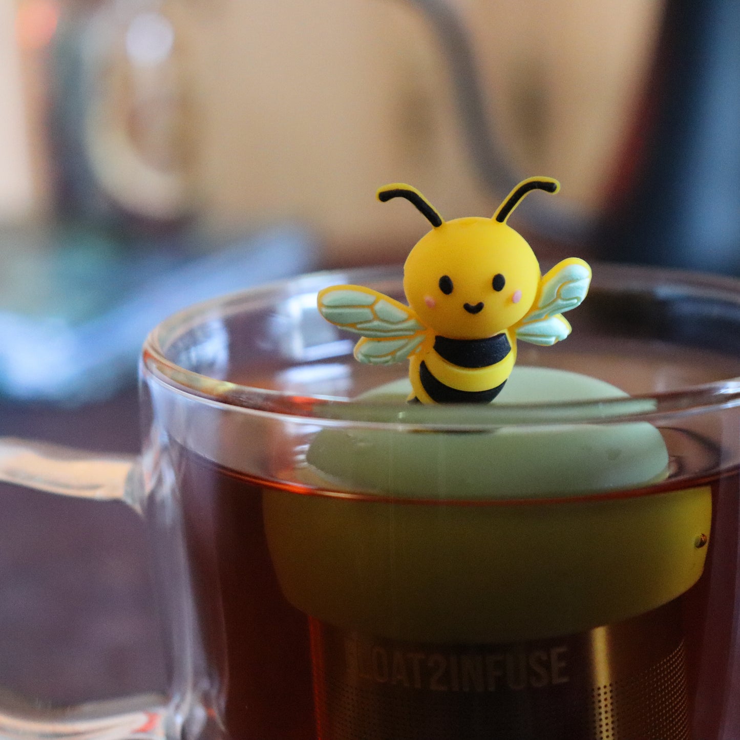 yellow and black bee animal tea infuser for loose leaf tea with a fine mesh stainless steel tea strainer that reads FLOAT2INFUSE  on a green base floating in a glass tea mug with tea kettle in the background
