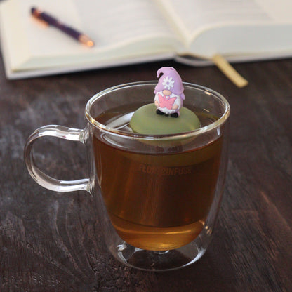 purple gnome holding pink butterfly tea infuser for loose leaf tea  with a fine mesh stainless steel tea infuser that reads FLOAT2INFUSE on a green base floating in a glass tea mug with notebook and pen in the background