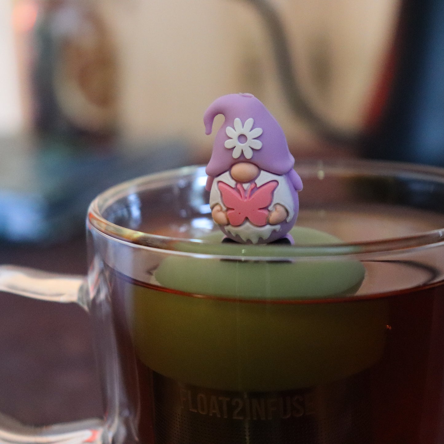 purple gnome holding pink butterfly tea infuser for loose leaf tea with a fine mesh stainless steel tea strainer that reads FLOAT2INFUSE  on a green base floating in a glass tea mug with tea kettle in the background