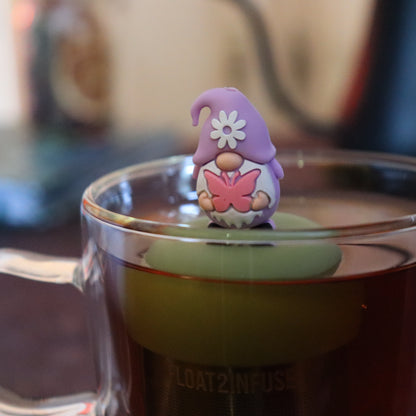 purple gnome holding pink butterfly tea infuser for loose leaf tea with a fine mesh stainless steel tea strainer that reads FLOAT2INFUSE  on a green base floating in a glass tea mug with tea kettle in the background
