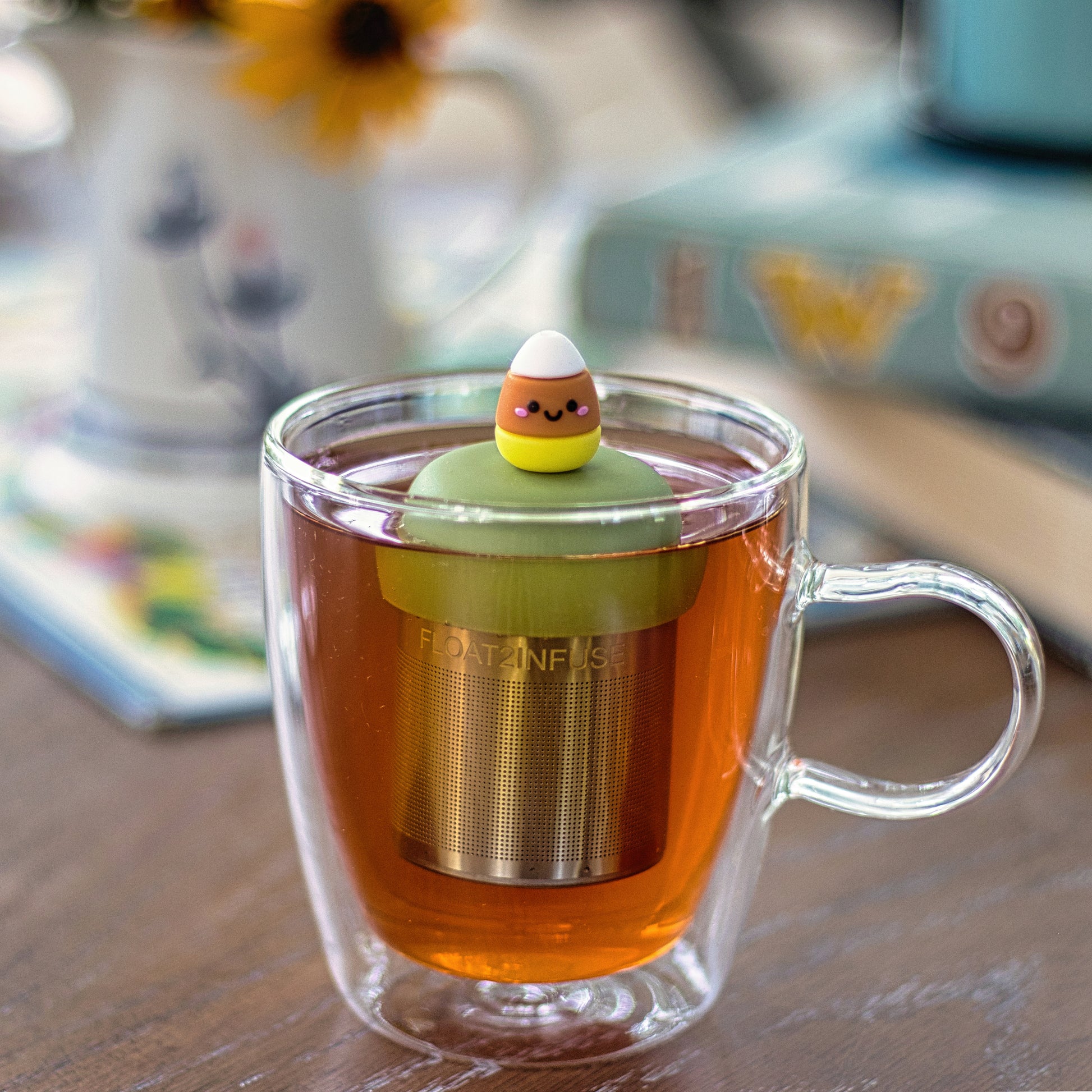 candy corn with a smiling face tea infuser for loose leaf tea with a fine mesh stainless steel tea strainer that reads FLOAT2INFUSE on a green base  floating in a glass tea mug 
with books in the background