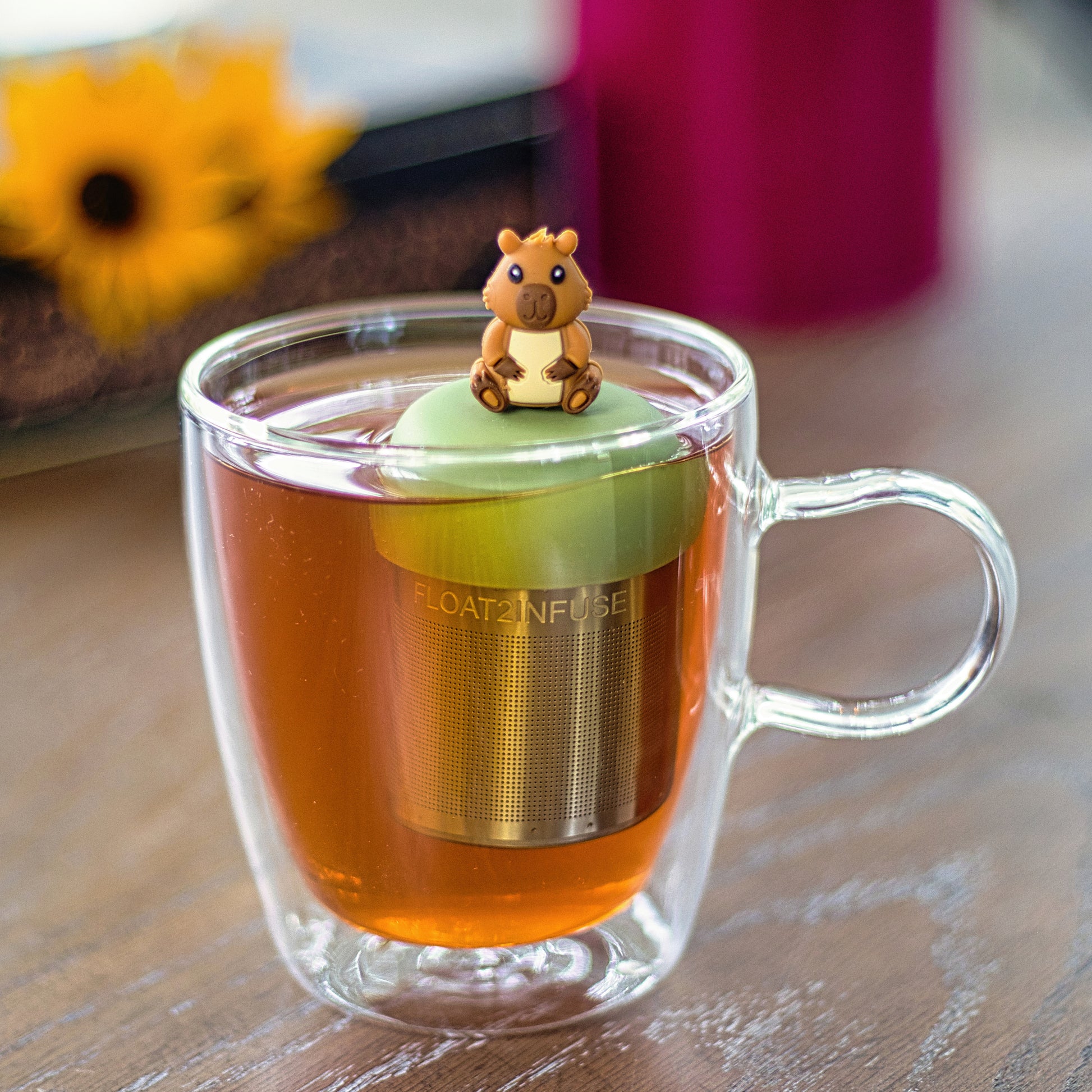happy brown capybara tea infuser for loose leaf tea with a fine mesh stainless steel tea strainer that reads FLOAT2INFUSE on a green base floating in a glass tea mug 
with flowers in the background