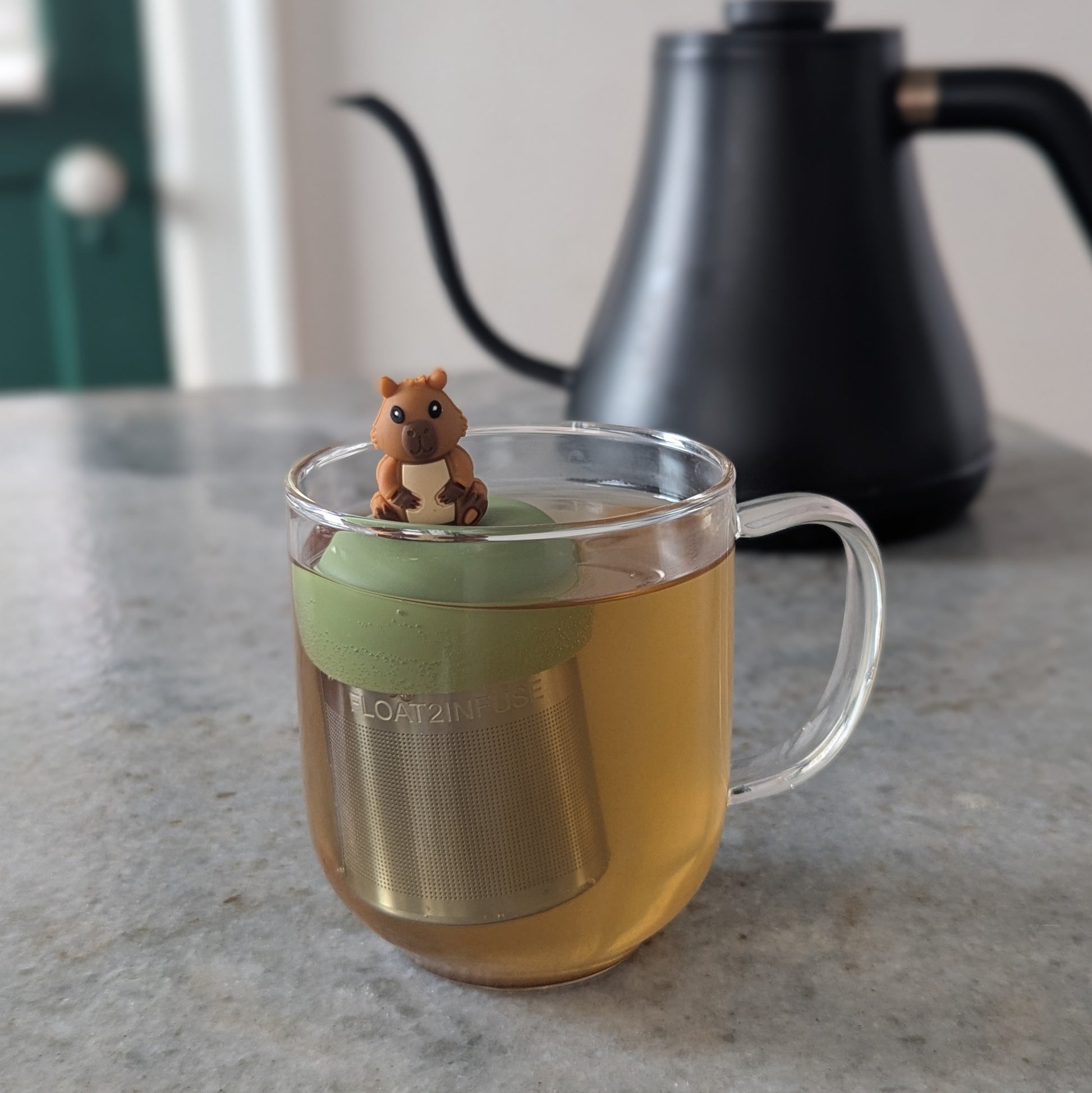 happy brown capybara tea infuser for loose leaf tea with a fine mesh stainless steel tea strainer that reads FLOAT2INFUSE on a green base floating in a glass tea mug 
with a tea kettle in the background