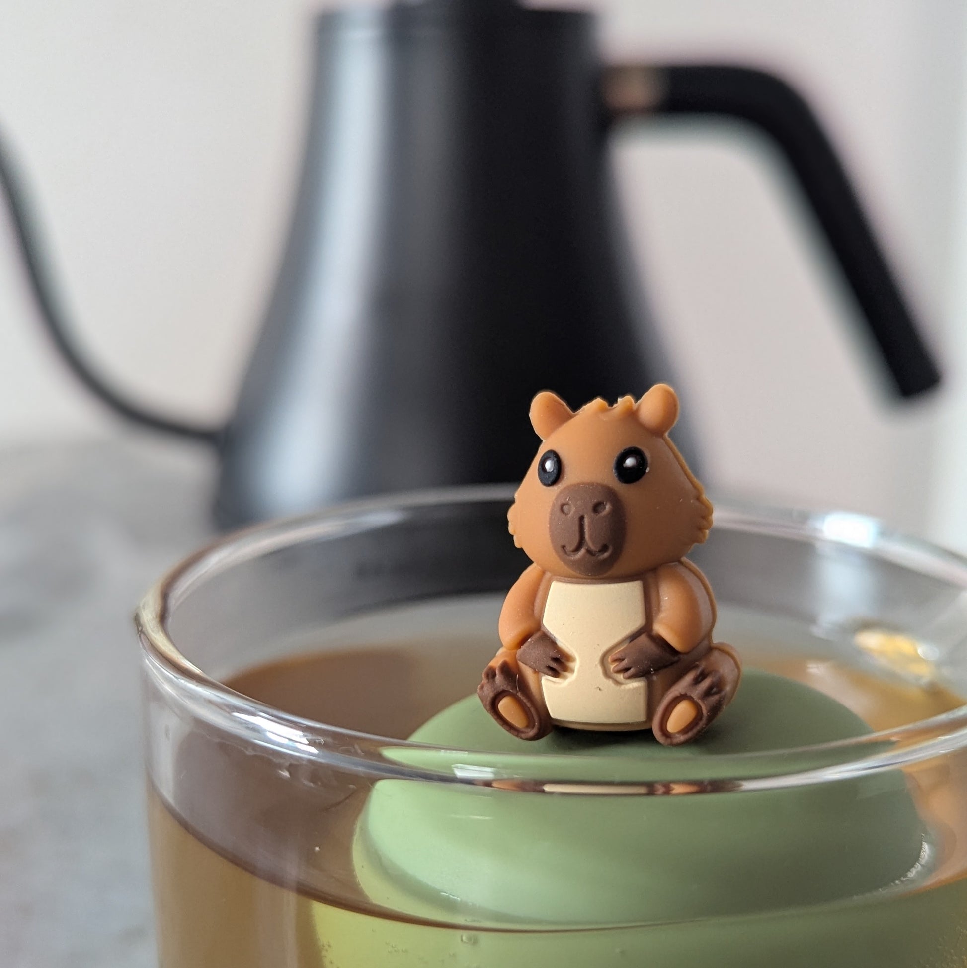 happy brown capybara tea infuser for loose leaf tea with a fine mesh stainless steel tea strainer that reads FLOAT2INFUSE on a green base floating in a glass tea mug 
with a tea kettle in the background