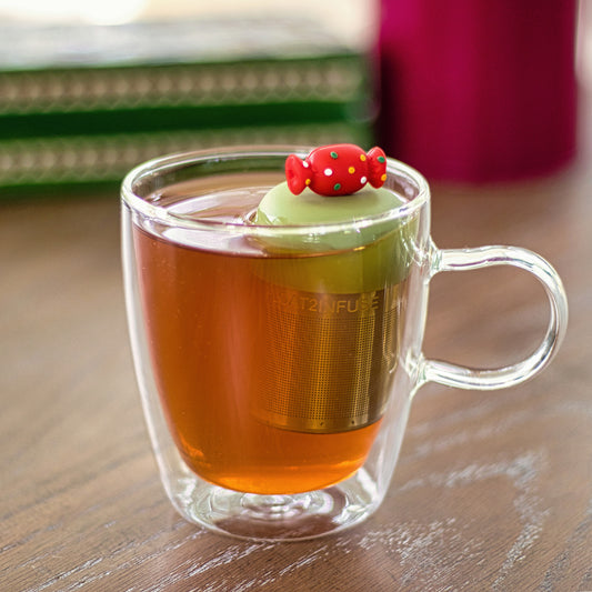 red holiday candy with green, yellow, and white polka dots tea infuser for loose leaf tea with a fine mesh stainless steel tea strainer that reads FLOAT2INFUSE on a green base  floating in a glass tea mug 
with books in the background