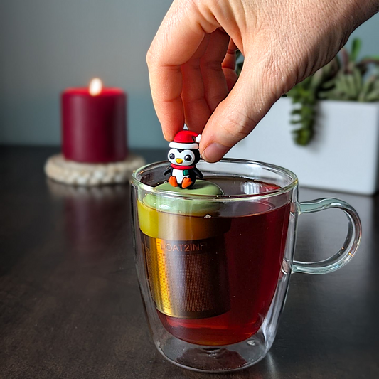 Black and white penguin wearing a red santa hat and a red and green scarf tea infuser for loose leaf tea with a fine mesh stainless steel tea strainer that reads FLOAT2INFUSE on a green base with a lit candle and plants in the background