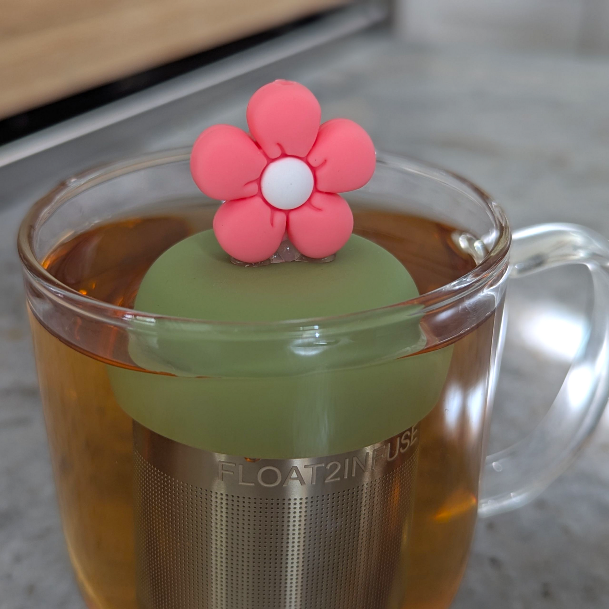 pink daisy with white center tea infuser for loose leaf tea with fine mesh stainless steel tea strainer that reads FLOAT2INFUSE on a green base floating in a glass tea mug