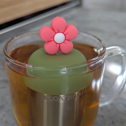 pink daisy with white center tea infuser for loose leaf tea with fine mesh stainless steel tea strainer that reads FLOAT2INFUSE on a green base floating in a glass tea mug