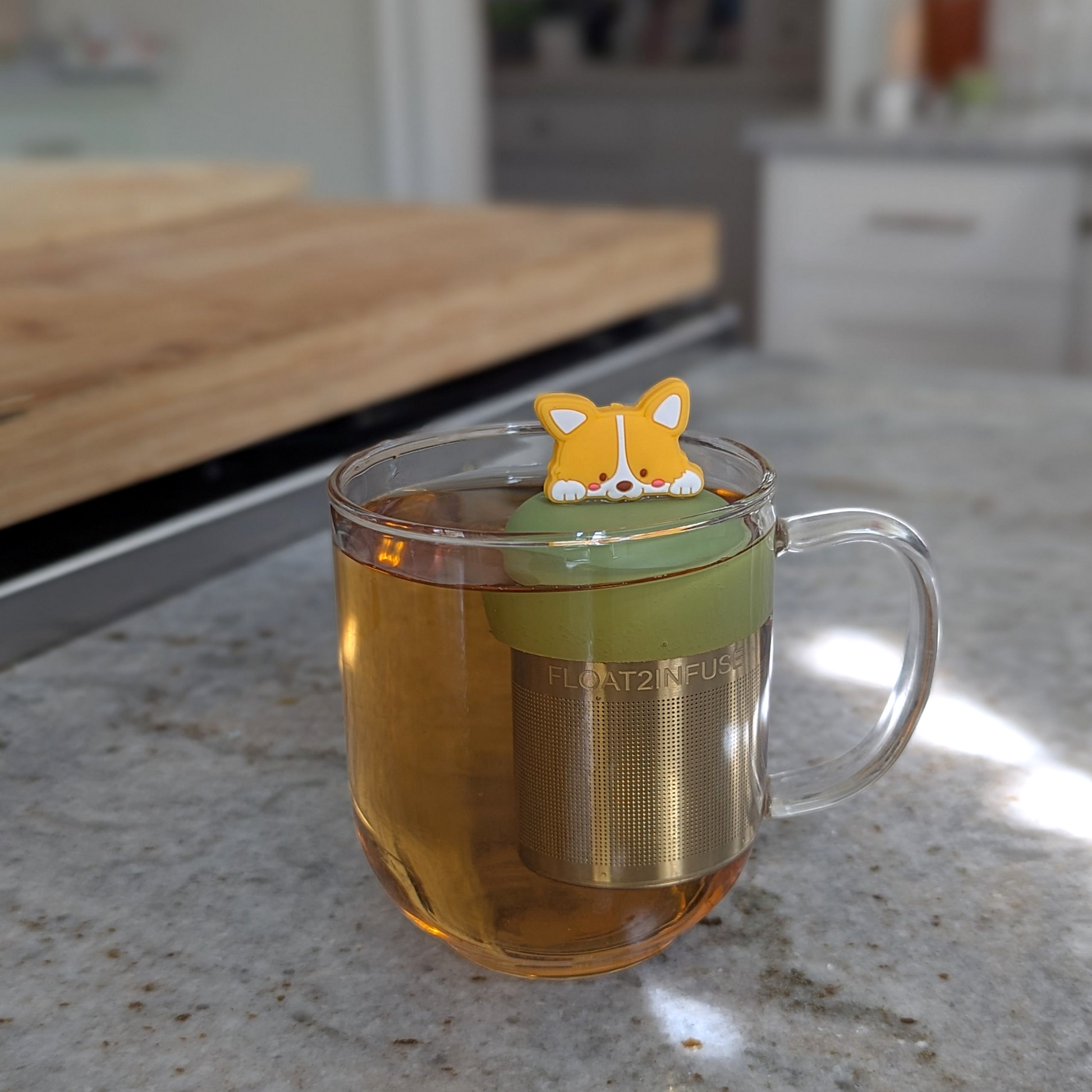smiling orange and white corgi puppy tea infuser for loose leaf tea with fine mesh stainless steel tea strainer that reads FLOAT2INFUSE on a green base floating in a glass tea mug with a kitchen counter in the background