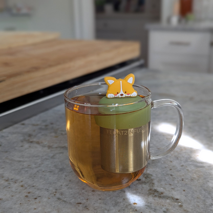 smiling orange and white corgi puppy tea infuser for loose leaf tea with fine mesh stainless steel tea strainer that reads FLOAT2INFUSE on a green base floating in a glass tea mug with a kitchen counter in the background