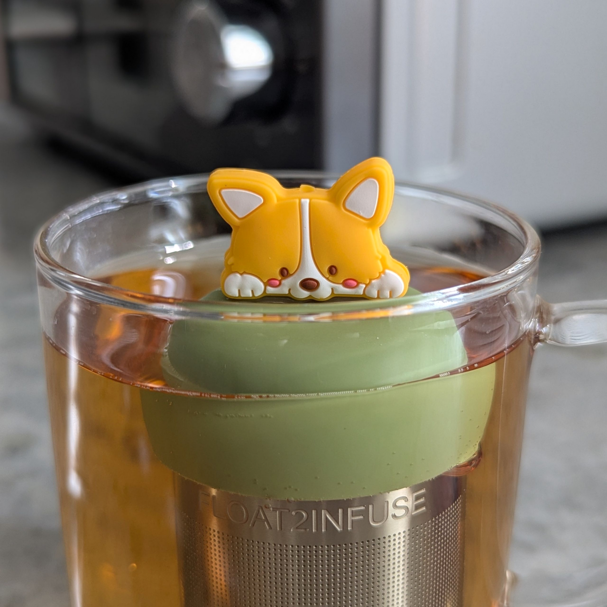 smiling orange and white corgi puppy tea infuser for loose leaf tea with fine mesh stainless steel tea strainer that reads FLOAT2INFUSE on a green base floating in a glass tea mug with a microwave in the background