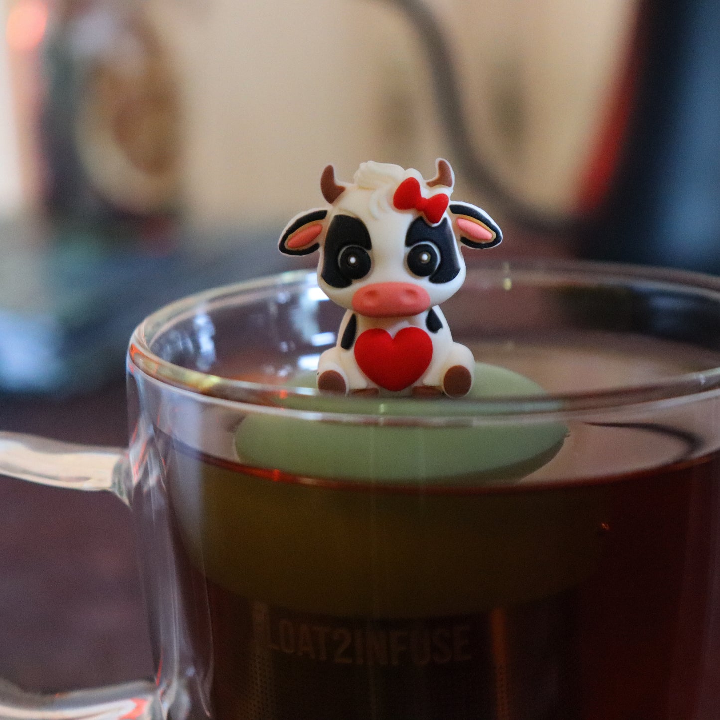 black and white cow with red bow and heart animal tea infuser for loose leaf tea  with a fine mesh stainless steel tea strainer that reads FLOAT2INFUSE on a green base floating in a glass tea mug with tea kettle in the background