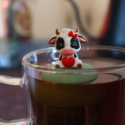 black and white cow with red bow and heart animal tea infuser for loose leaf tea  with a fine mesh stainless steel tea strainer that reads FLOAT2INFUSE on a green base floating in a glass tea mug with tea kettle in the background