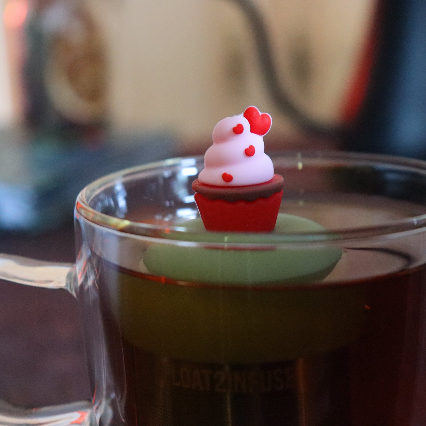 red and pink cupcake with hearts tea infuser for loose leaf tea with a fine mesh stainless steel tea strainer that reads FLOAT2INFUSE on a green base  floating in a glass tea mug with tea kettle in the background