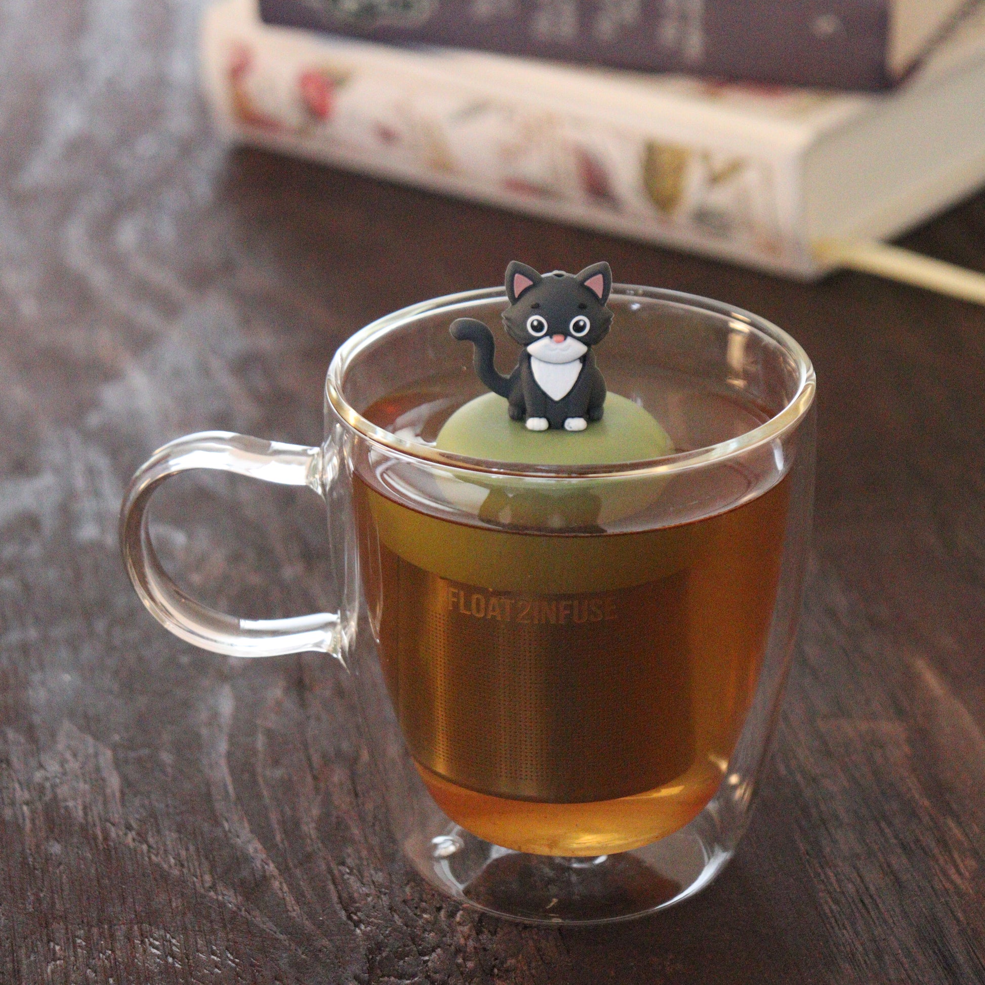 grey and white cat animal tea infuser for loose leaf tea on a green base with a fine mesh stainless steel tea infuser that reads FLOAT2INFUSE floating in a glass tea mug with books in the background