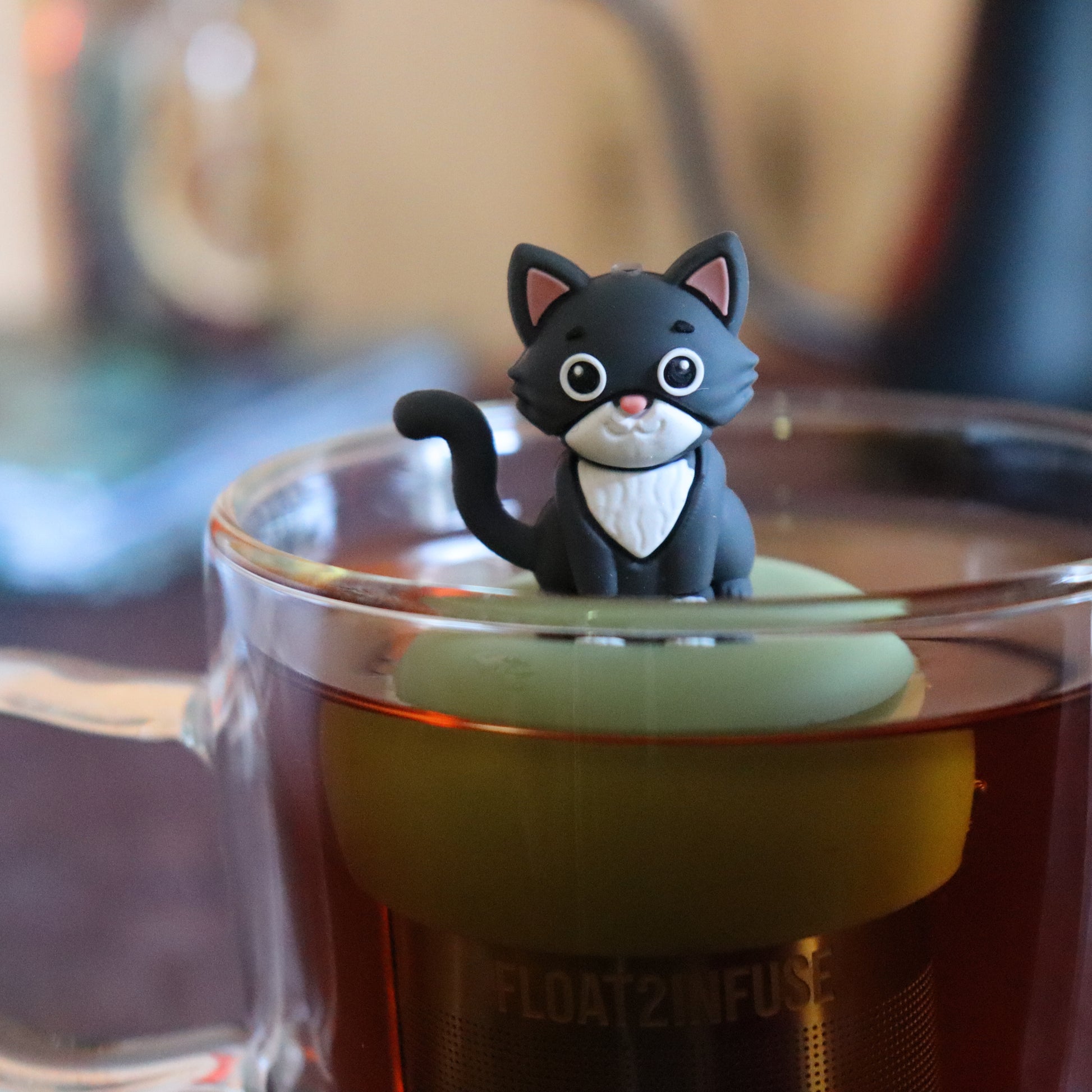 grey and white cat animal tea infuser for loose leaf tea on a green base with a fine mesh stainless steel tea strainer that reads FLOAT2INFUSE floating in a glass tea mug with tea kettle in the background