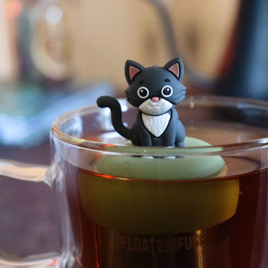 grey and white cat animal tea infuser for loose leaf tea on a green base with a fine mesh stainless steel tea strainer that reads FLOAT2INFUSE floating in a glass tea mug with tea kettle in the background