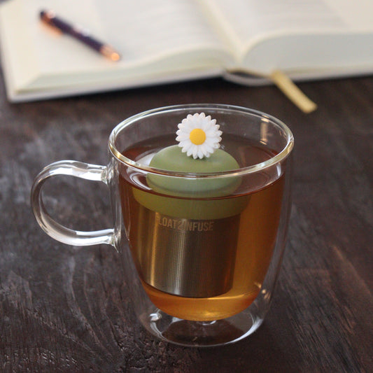 white daisy tea infuser for loose leaf tea on a green base with a fine mesh stainless steel tea infuser that reads FLOAT2INFUSE floating in a glass tea mug with notebook and pen in the background