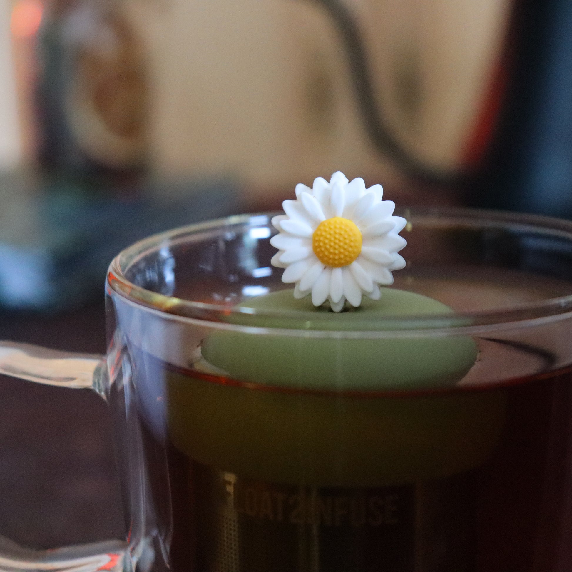 white daisy tea infuser for loose leaf tea  with a fine mesh stainless steel tea strainer that reads FLOAT2INFUSE on a green base floating in a glass tea mug with tea kettle in the background