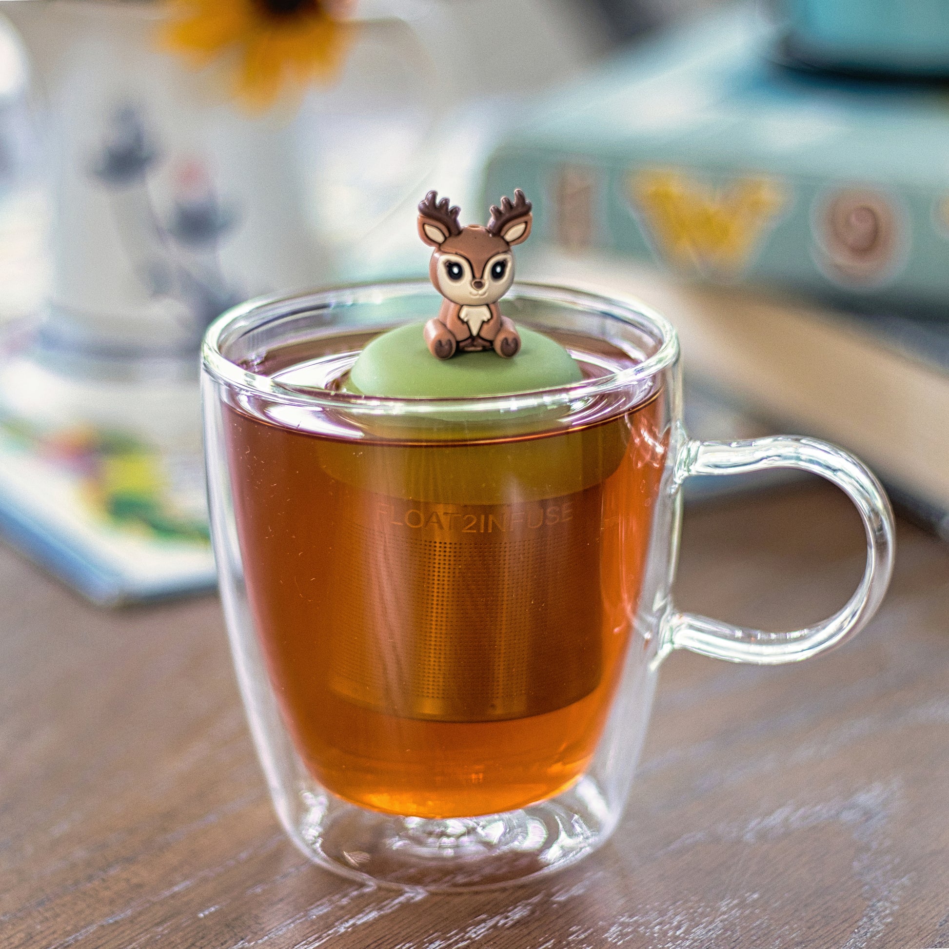 happy brown and beige deer tea infuser for loose leaf tea with a fine mesh stainless steel tea strainer that reads FLOAT2INFUSE on a green base floating in a glass tea mug 
with books in the background