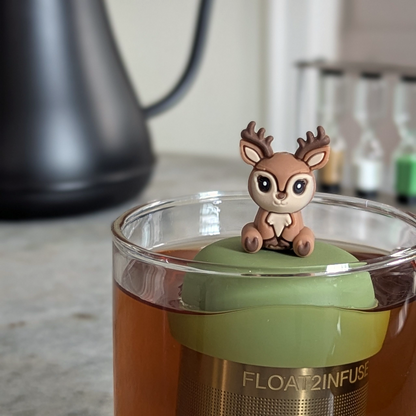 happy brown and beige deer tea infuser for loose leaf tea with a fine mesh stainless steel tea strainer that reads FLOAT2INFUSE on a green base floating in a glass tea mug 
with a tea kettle in the background