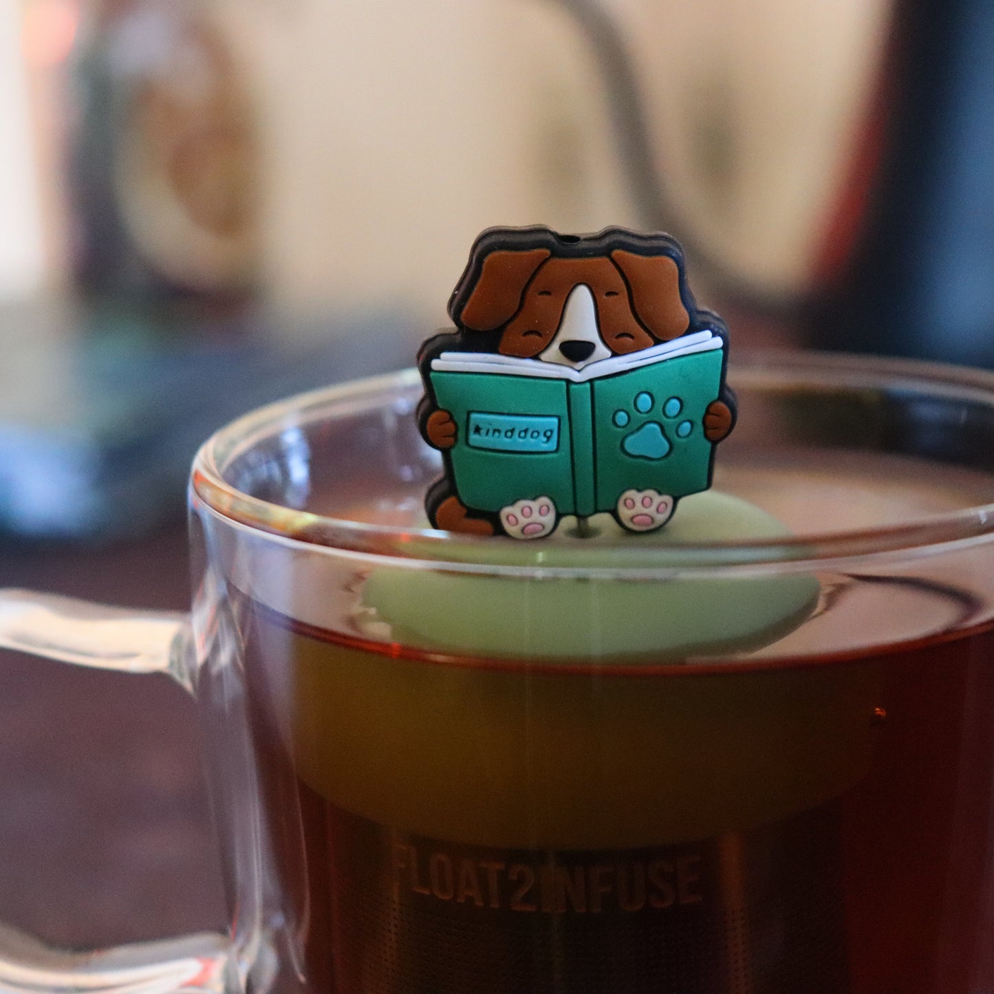 brown dog reading green book animal tea infuser for loose leaf tea  with a fine mesh stainless steel tea strainer that reads FLOAT2INFUSE on a green base floating in a glass tea mug with tea kettle in the background