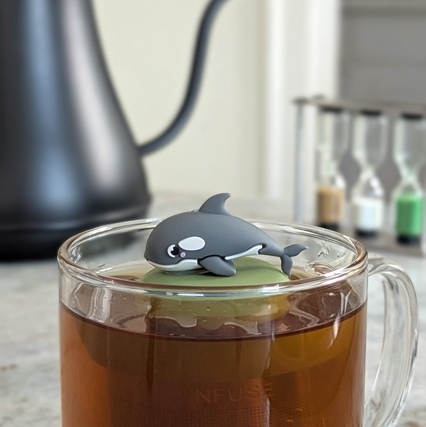 blushing grey dolphin tea infuser for loose leaf tea  with a fine mesh stainless steel strainer that reads FLOAT2INFUSE on a green base floating in a glass tea mug with a tea kettle in the background